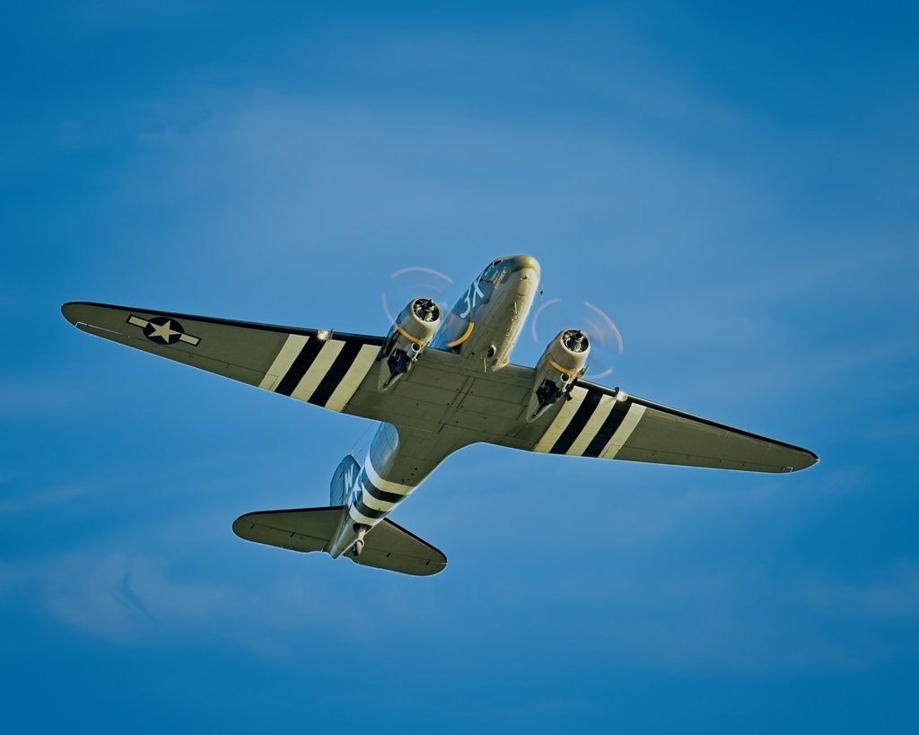 C-47 That’s All, Brother flying overhead with D-Day invasion stripes visible beneath its wings and fuselage.